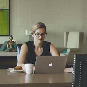 The Career Academy Stay Motivated - Young Professional Woman sitting at desk with muffin and coffee working on laptop